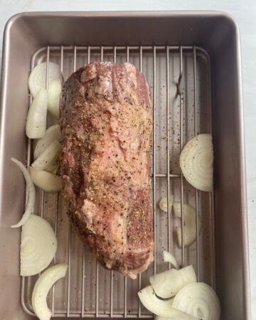 An eye round roast in a pan seasoned with Kosher salt and black pepper with sliced onions. 