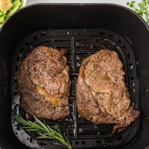 Air fryer basket with rib eye steaks that have been cooked.