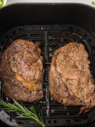 Air fryer basket with rib eye steaks that have been cooked.