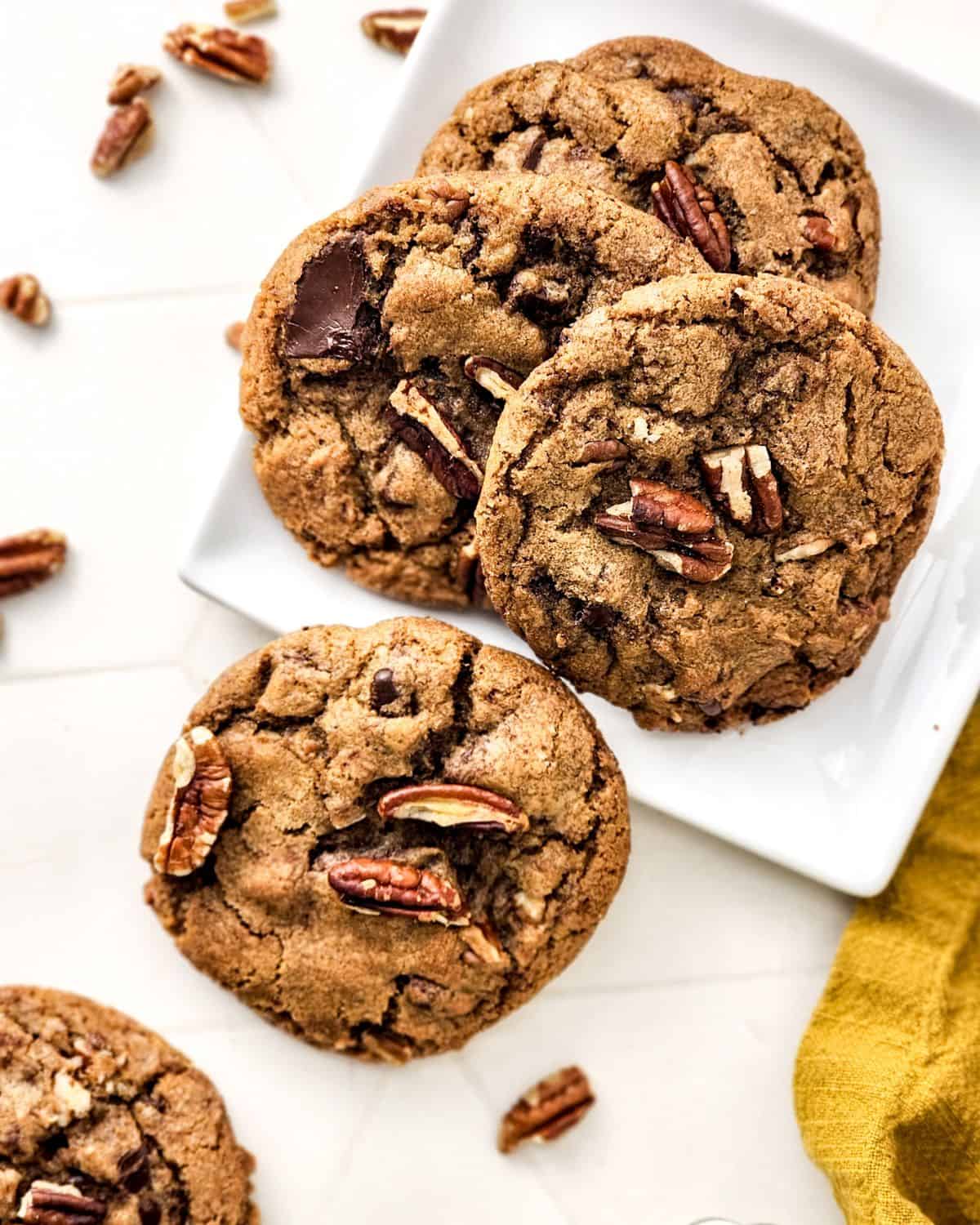 Brown butter pecan cookies with chocolate chips on a plate with pecans all around the plate. 