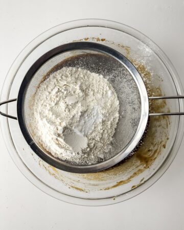 A glass bowl of cookie batter with a sifter over the bowl with flour and salt being sifted into the batter.