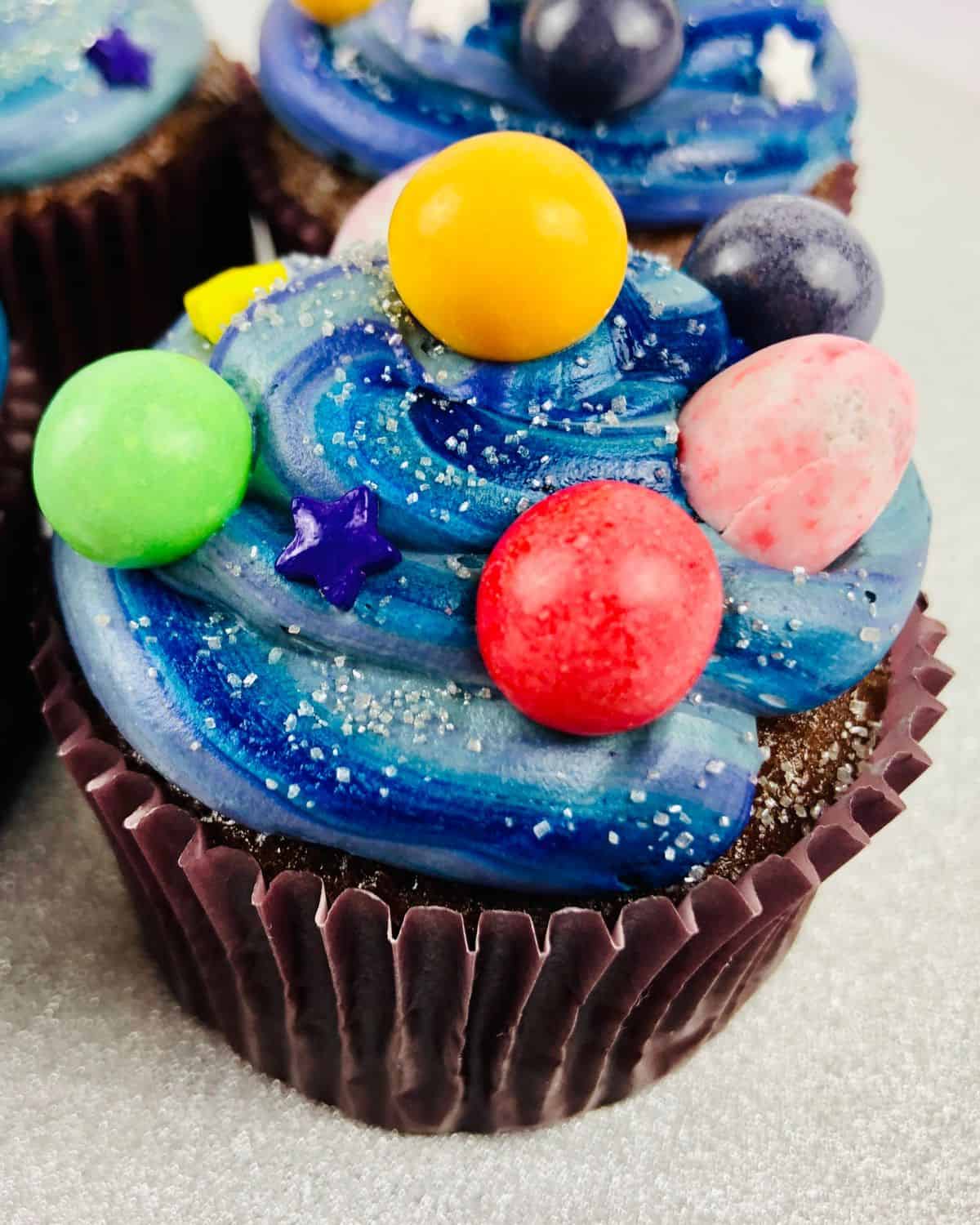 A cupcake topped with various blue colored frosting and round candies as the planets for galaxy cupcakes.