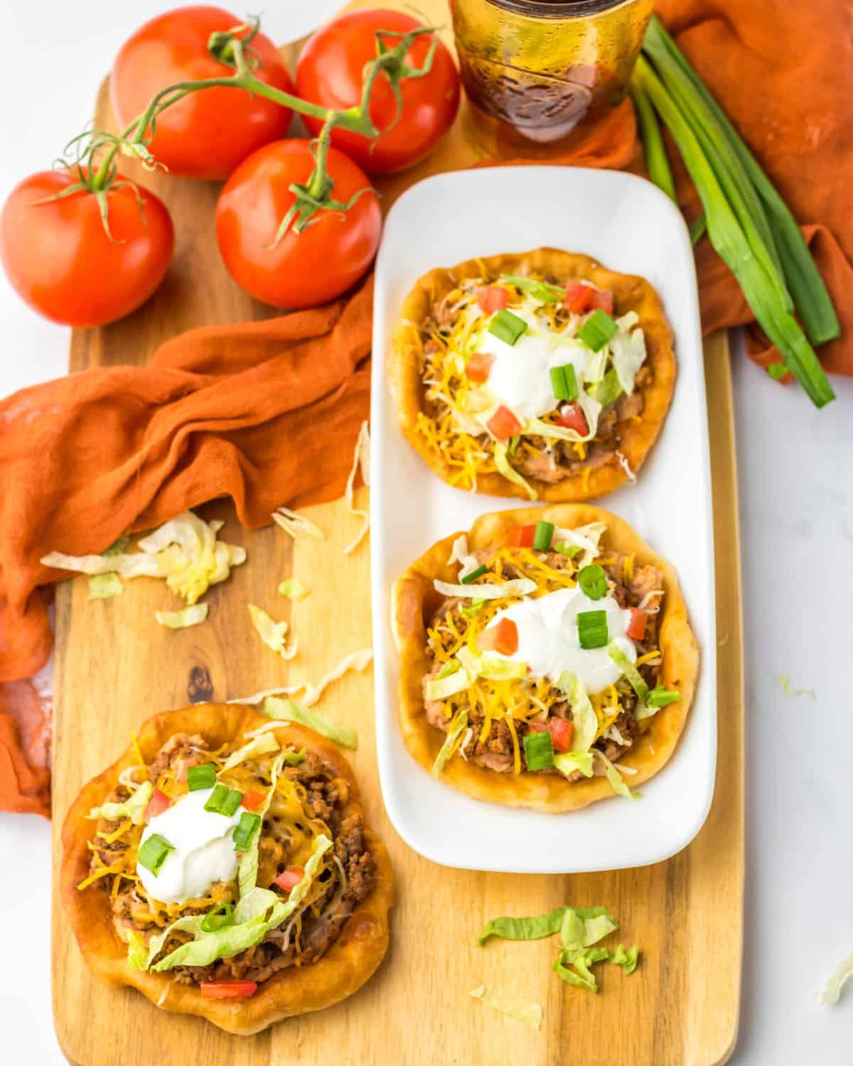 A wooden board with Indian Fry Bread Tacos with taco meat, lettuce, tomato.