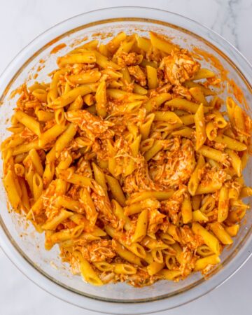 A glass mixing bowl with cooked pasta and chicken with marinara sauce to make a chicken parm casserole with pasta.