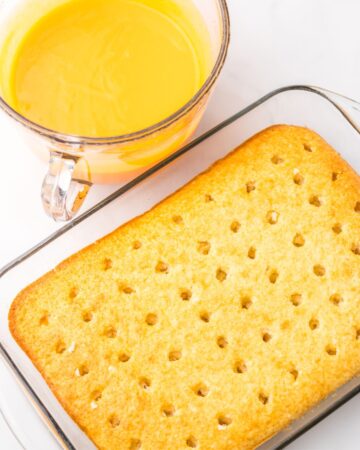 A baked cake with poke holes in it and a glass measuring cup with orange gelatin and whipped cream to make a cream topping for poke cake.