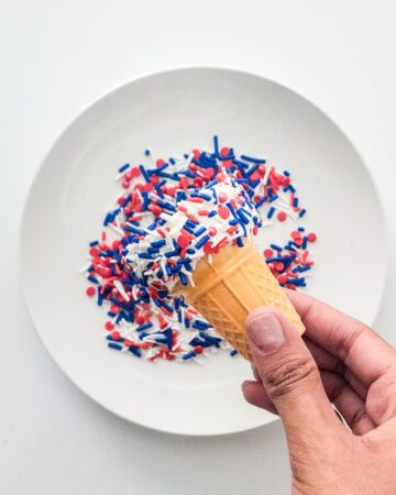 No bake cheesecake waffle cones being dipped into red white blue sprinkles.