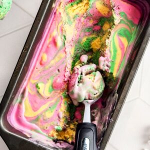 Homemade rainbow sherbet in a loaf pan being picked up with a scoop.