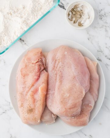 A glass dish with a chicken cutlet seasoned with salt, black pepper, sitting in flour.