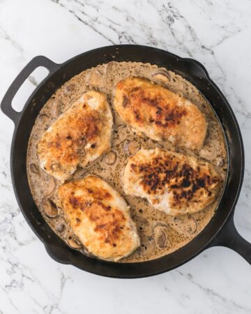 A cast iron pan with cooked season chicken breasts in a creamy marsala sauce with mushrooms.