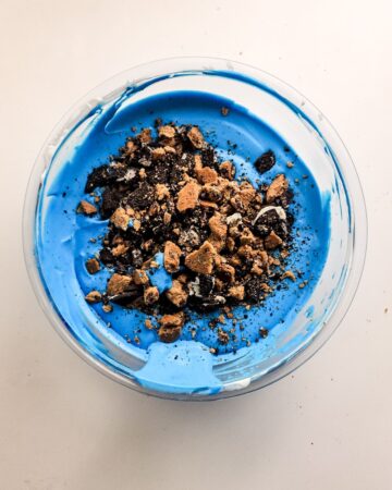A glass bowl with blue ice cream and chopped up cookies to make homemade cookie monster ice cream.