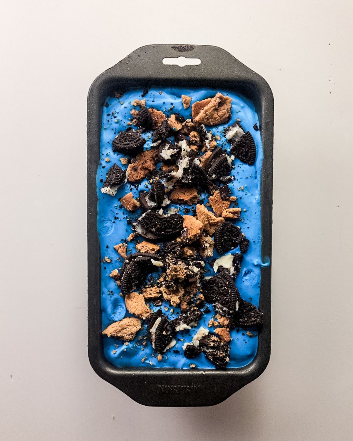 A loaf pan with no churn cookie monster ice cream topped with chopped cookies.