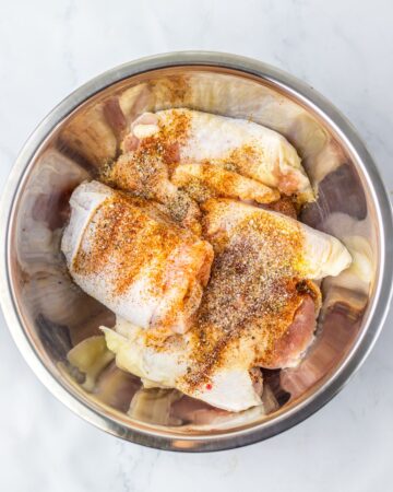 A stainless steel bowl with skin on chicken thighs with seasoning sprinkled over the thighs. 