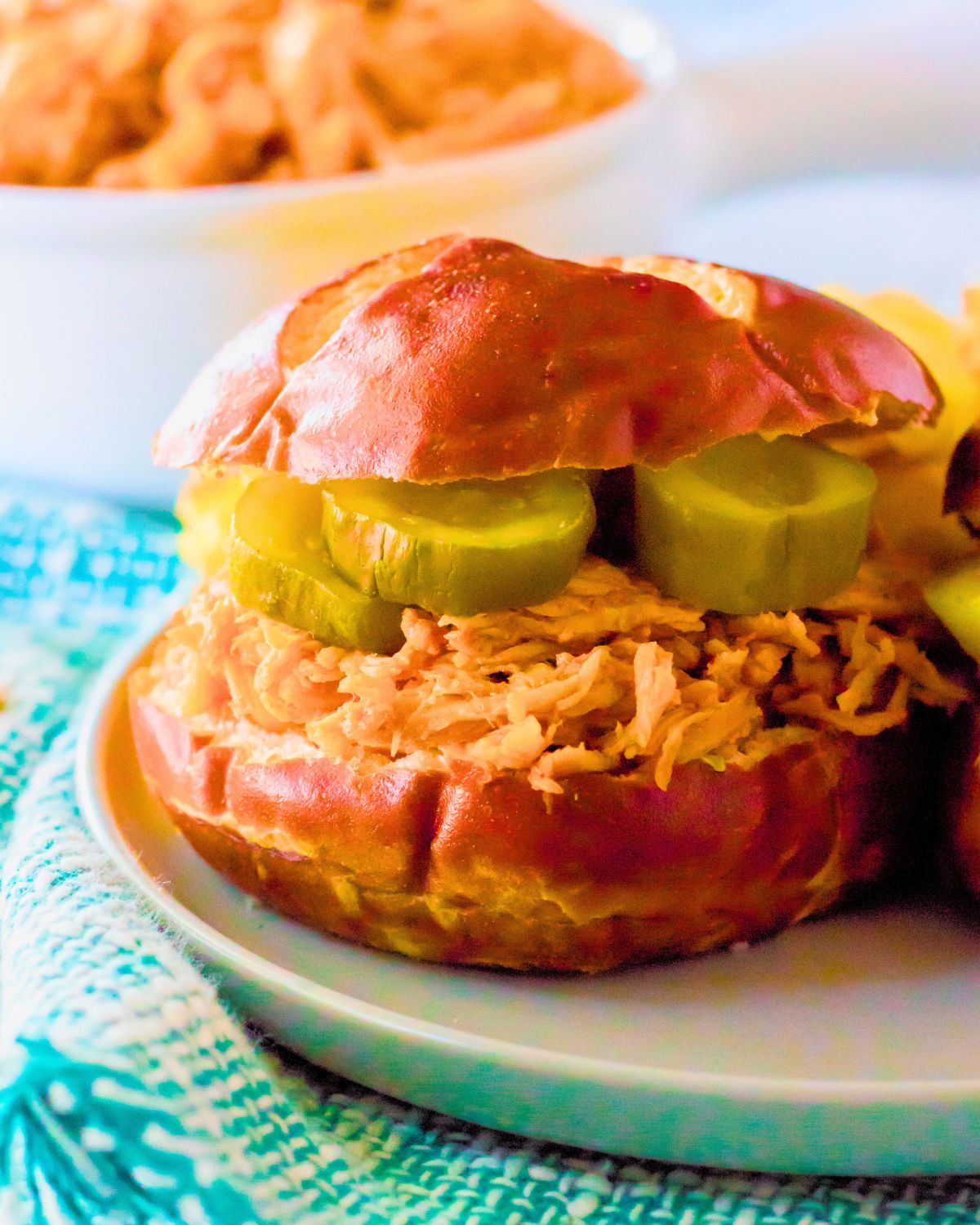 White plate with a brioche bun sliced and topped with hot honey shredded chicken and pickles