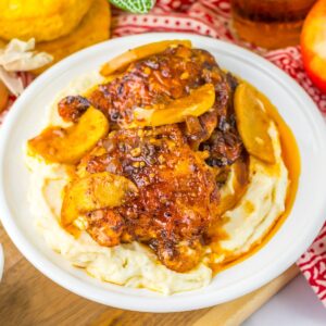 A plate with mashed potatoes topped with chicken thighs and apples in an apple cider sauce.