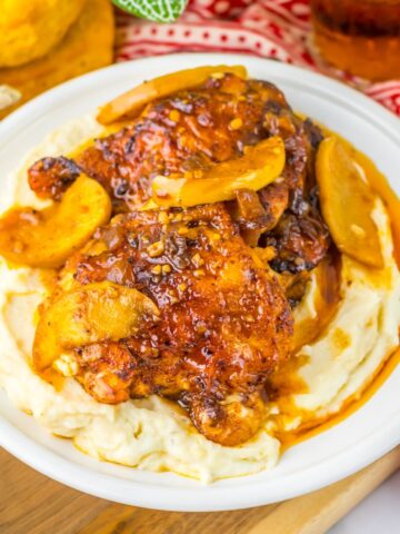 A plate with mashed potatoes topped with chicken thighs and apples in an apple cider sauce.