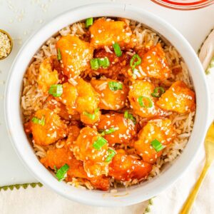 Orange chicken in a bowl with rice and topped with sesame seeds and chopped green onion.