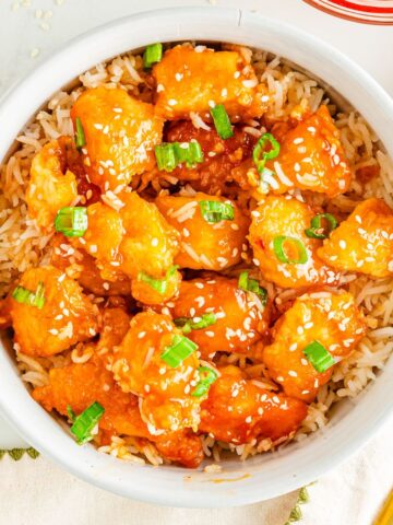 Orange chicken in a bowl with rice and topped with sesame seeds and chopped green onion.