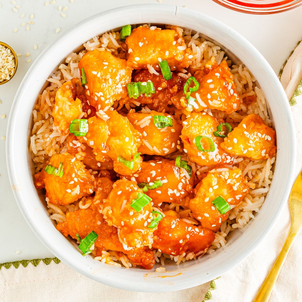Orange chicken in a bowl with rice and topped with sesame seeds and chopped green onion.