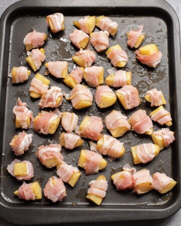 Potato wedges wrapped in bacon on a baking sheet.