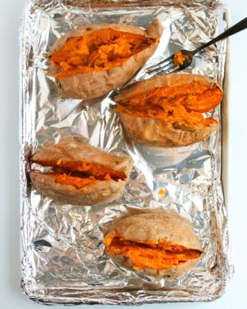 A baking sheet lined with aluminum foil with baked potatoes sliced open.