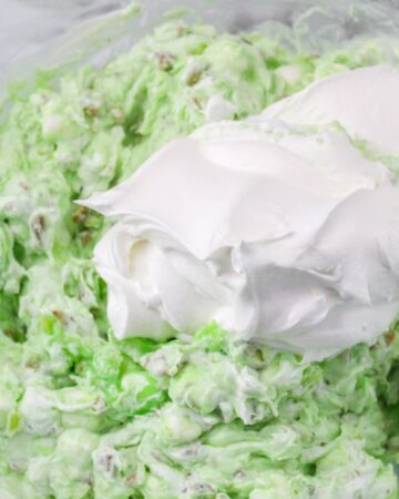 Pistachio pudding, marshmallows, pecans in a bowl with whipped topping being folded in to make Watergate Salad.