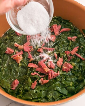 Sugar being poured over Collard greens with sliced ham.