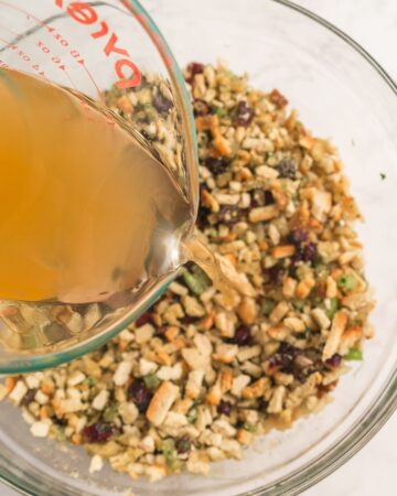 A bowl of stuffing with cranberries and a glass measuring cup with stock being poured into the bowl. 