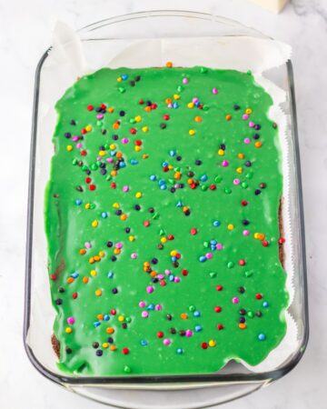 A baking pan with brownies topped with green chocolate ganache and colored sprinkles to make cosmic brownies with christmas tree shapes.