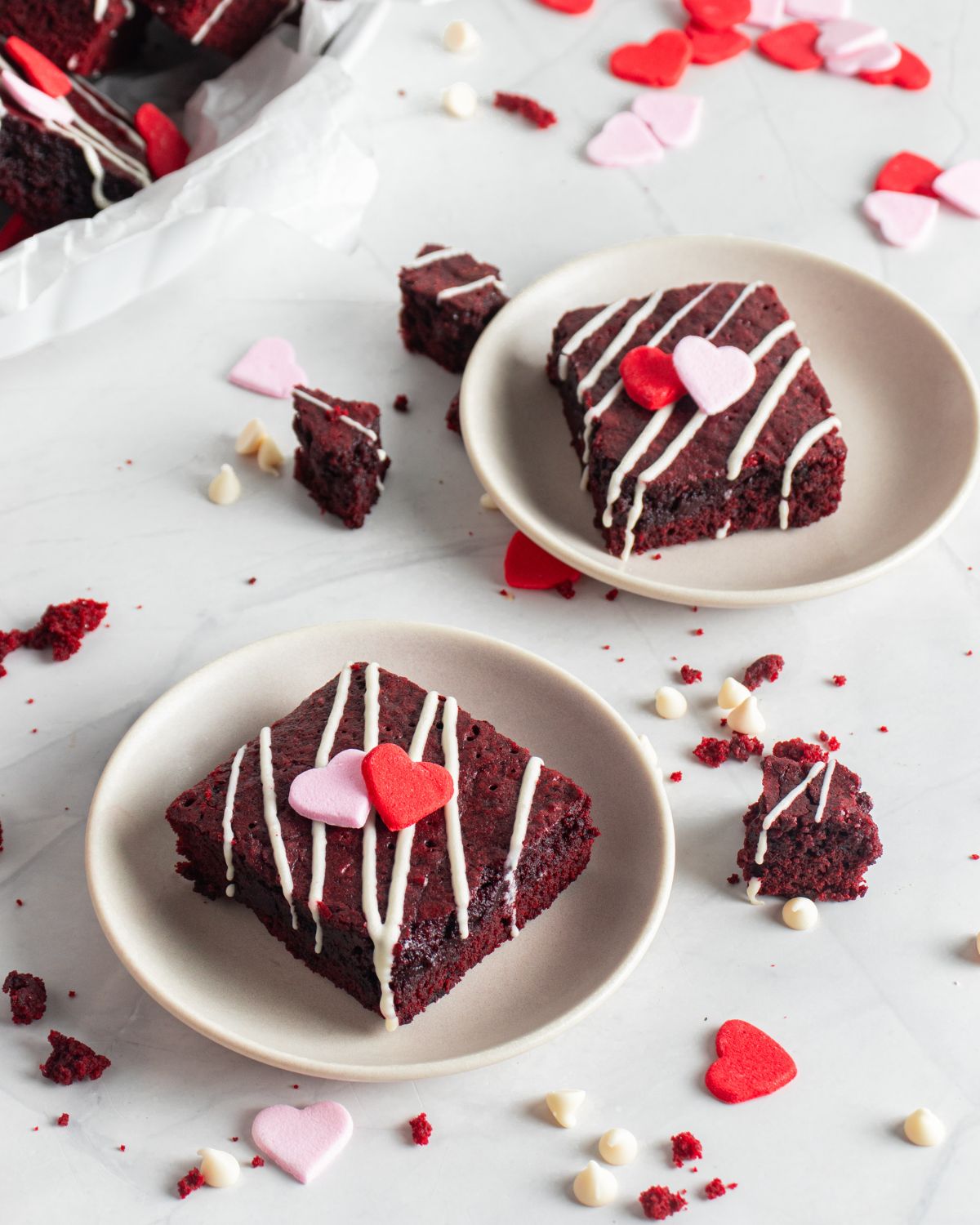 Two plates with brownie squares that are red velvet brownies topped with a white chocolate drizzle.