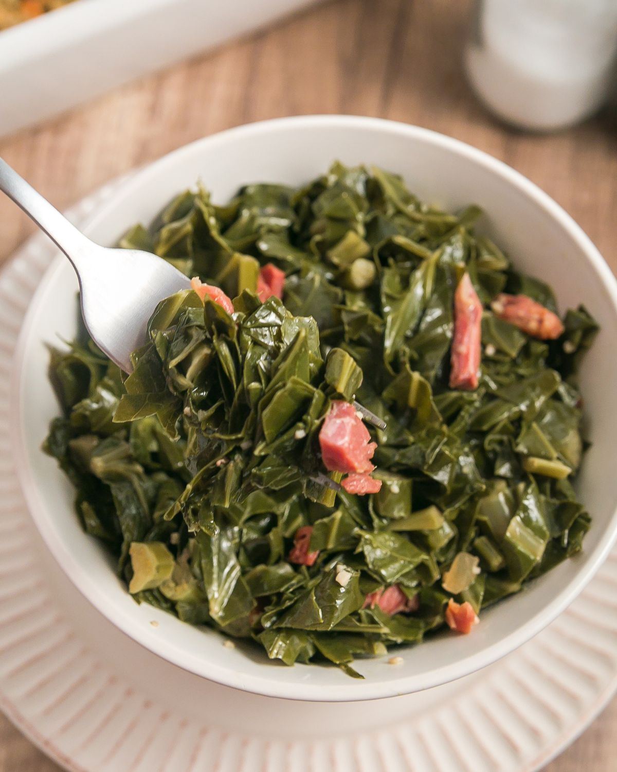 A white bowl with cooked collard greens and ham hocks being picked up with a fork.