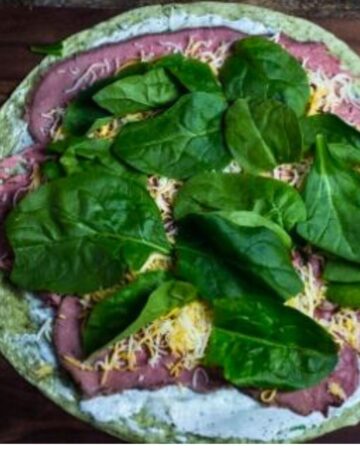 Roast beef tortilla rollup being prepared with roast beef, cheddar cheese, and baby spinach. 