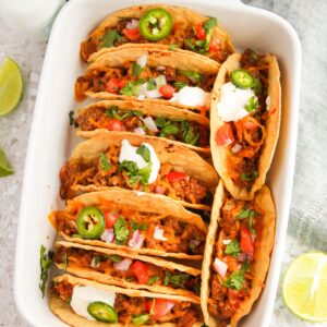 A white baking dish with baked beef tacos in hard shell tacos topped with chopped tomatoes, cilantro, jalapeno, and sour cream.