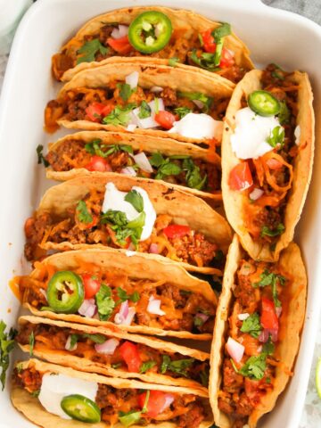 A white baking dish with baked beef tacos in hard shell tacos topped with chopped tomatoes, cilantro, jalapeno, and sour cream.