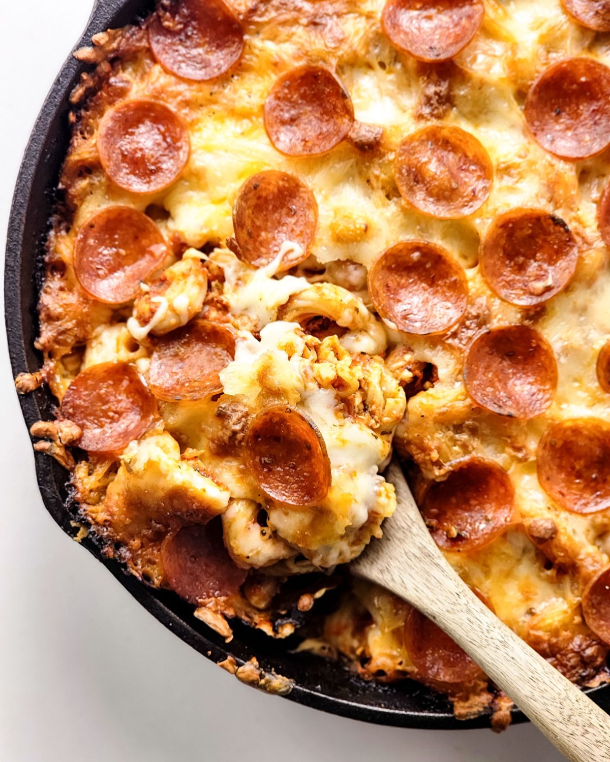 A wooden spoon picking up a scoop of pasta with marinara and cheese topped with pepperoni.