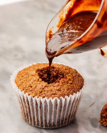 A Baileys cupcake with a portion taken out to have chocolate syrup being poured into the center of the cupcake. 