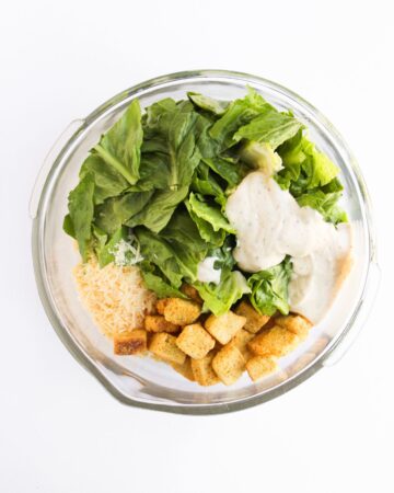 A glass mixing bowl with chopped romaine lettuce, parmesan cheese, croutons, caesar dressing with rotini pasta to make Chicken Caesar Pasta salad.