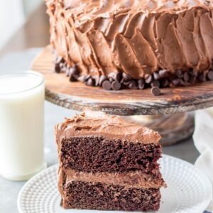 Chocolate cake slice with the cake on a cake stand behind it.