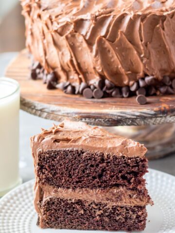 Chocolate cake slice with the cake on a cake stand behind it.