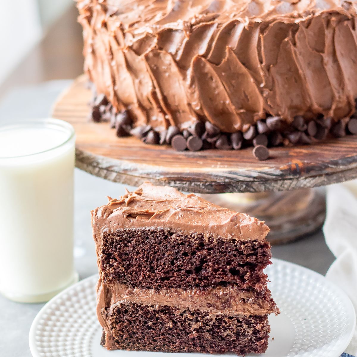 Chocolate cake slice with the cake on a cake stand behind it.