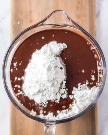 A glass measuring cup with ingredients to make chocolate frosting for chocolate cake. 