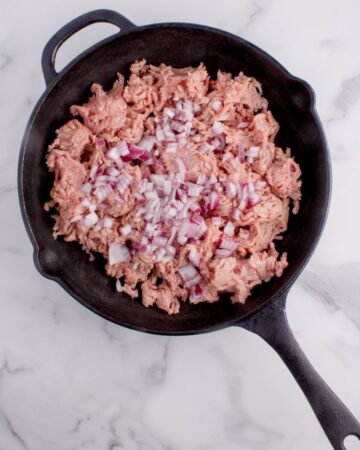 A cast iron pan with ground beef and diced onion to make hamburger hash brown casserole.