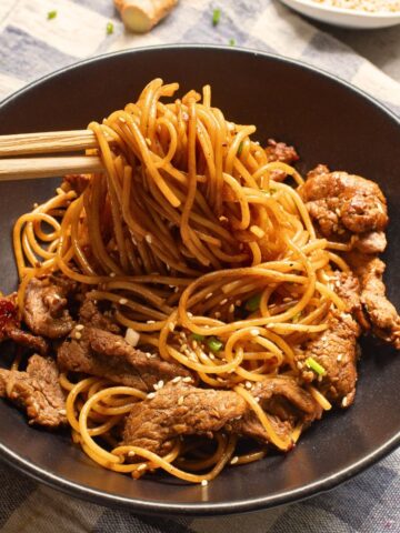 A black plate with thinly sliced cooked steak and noodles for a hibachi steak and noodles recipe topped with sesame seeds and chop sticks picking up the noodles.