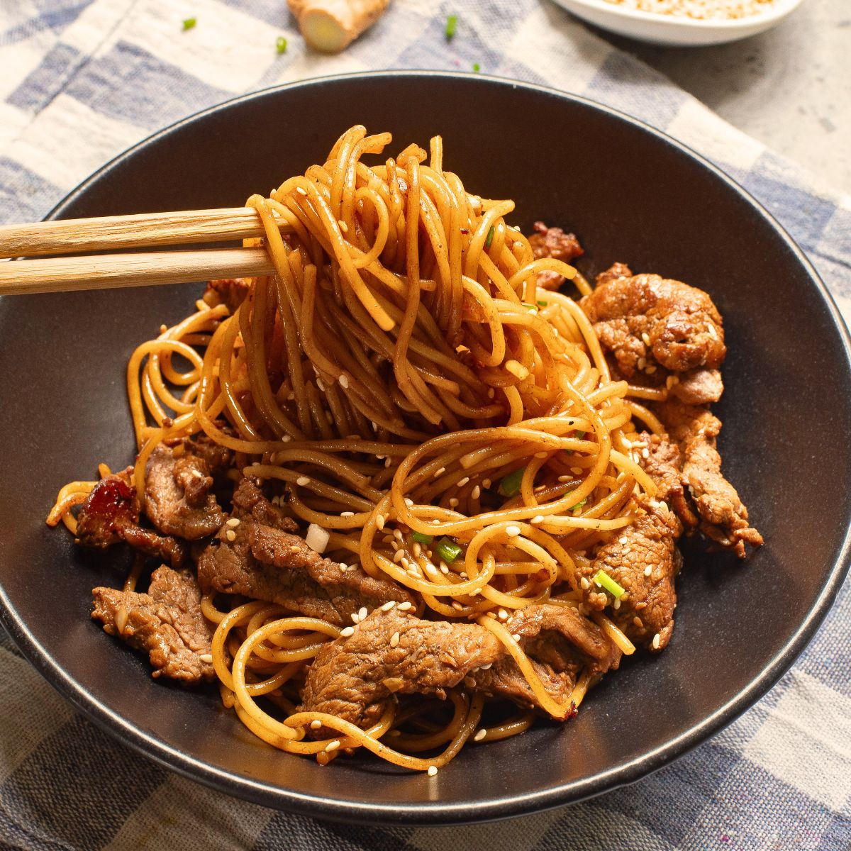 A black plate with thinly sliced cooked steak and noodles for a hibachi steak and noodles recipe topped with sesame seeds and chop sticks picking up the noodles.