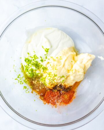 A large glass cowl with mayonnaise, lime zest and seasoning to make chicken corn salad.