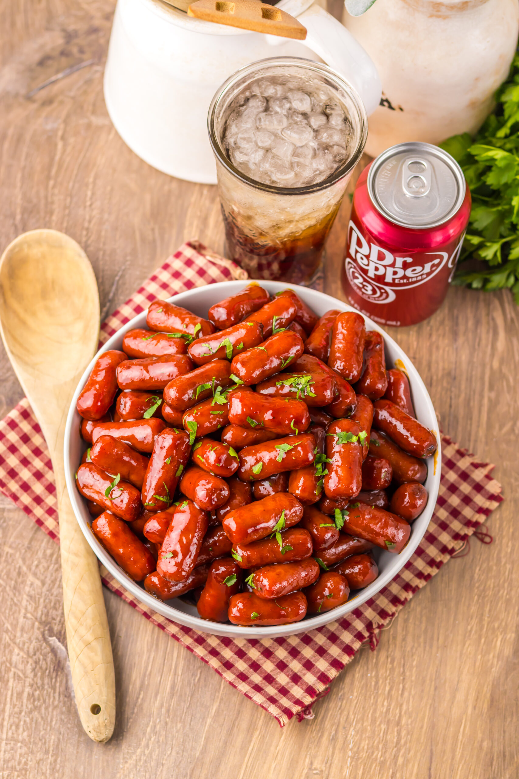 A white bowl with lil smokies in the bowl sitting on a checkered red and tan linen with Dr Pepper in a glass behind the bowl.