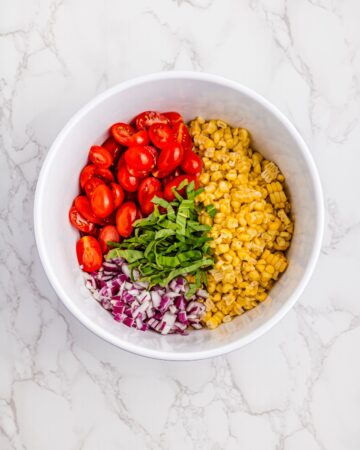 A white bowl with ingredients for tomato, corn, red onion, and herbs.