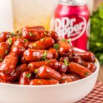 A white bowl with little smokies cooked in Dr Pepper and BBQ sauce on a checkered linen. A can of Dr Pepper in the background.