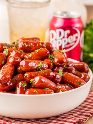 A white bowl with little smokies cooked in Dr Pepper and BBQ sauce on a checkered linen. A can of Dr Pepper in the background.