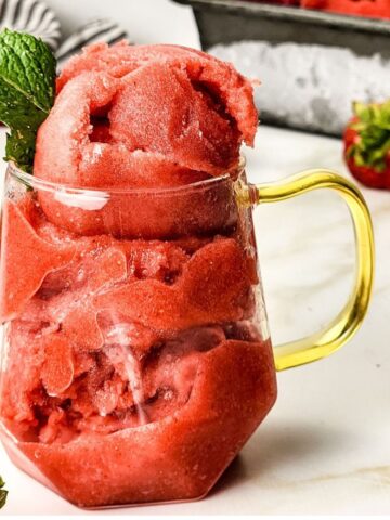 A glass cup with a gold handle with scoops of strawberry sorbet in the cup.