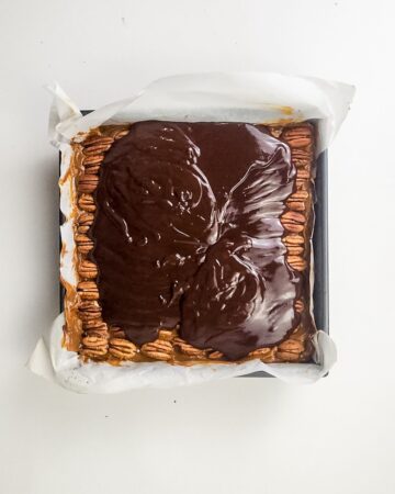 An 8 x 8 inch baking pan with the layers to make millionaire brownies - with the chocolate ganache above whole pecans.